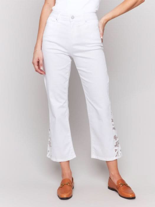 A woman in a white sleeveless top and Charlie B's Lace Cut Side Bootcut Twill Pants with embroidered details stands against a plain background, wearing brown shoes.