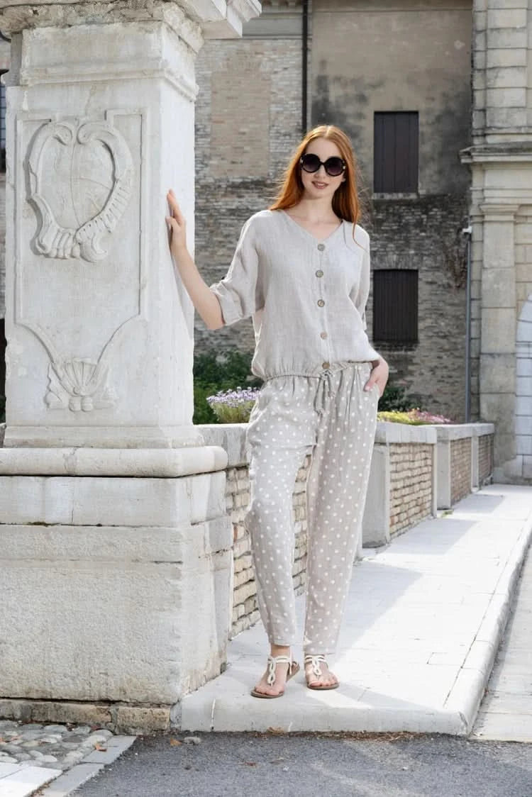 A woman stands by a stone pillar in the sun, wearing sunglasses, a light button-up shirt, and Variations Polka Dot Slim Pant.
