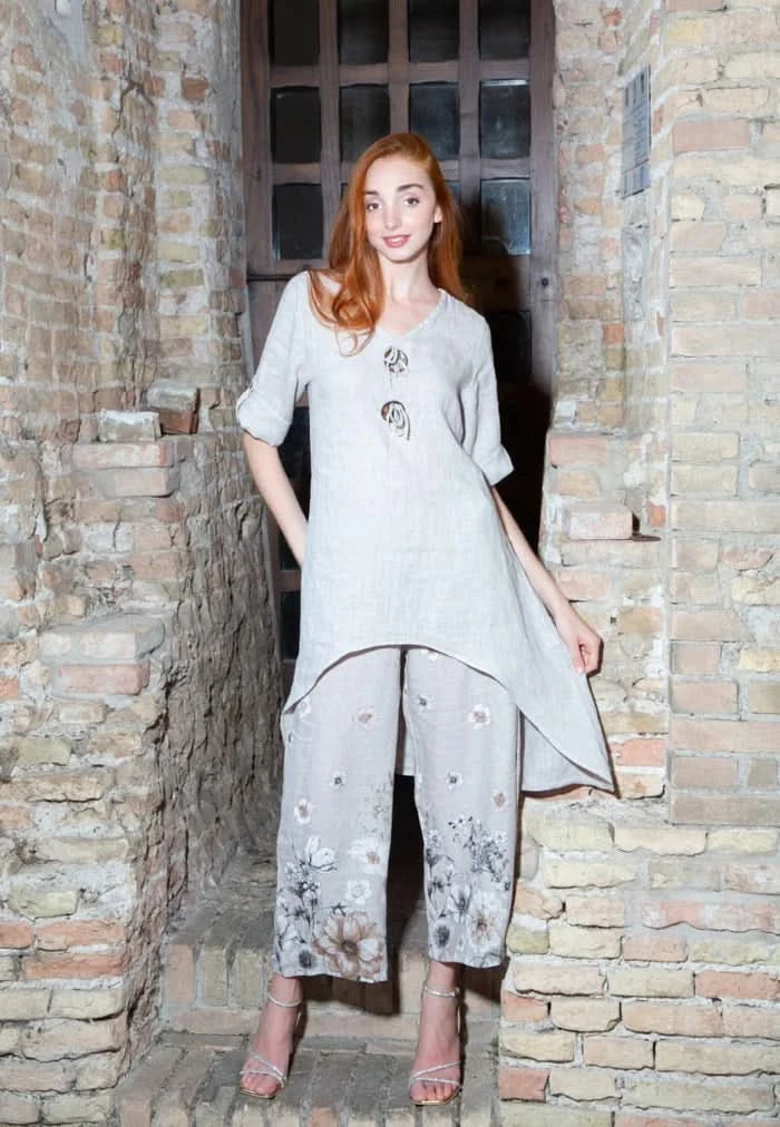 A woman with long red hair stands before a brick wall, wearing the Variations Linen Tunic Top with Toggle Closures and floral wide-leg pants, paired with heeled sandals.