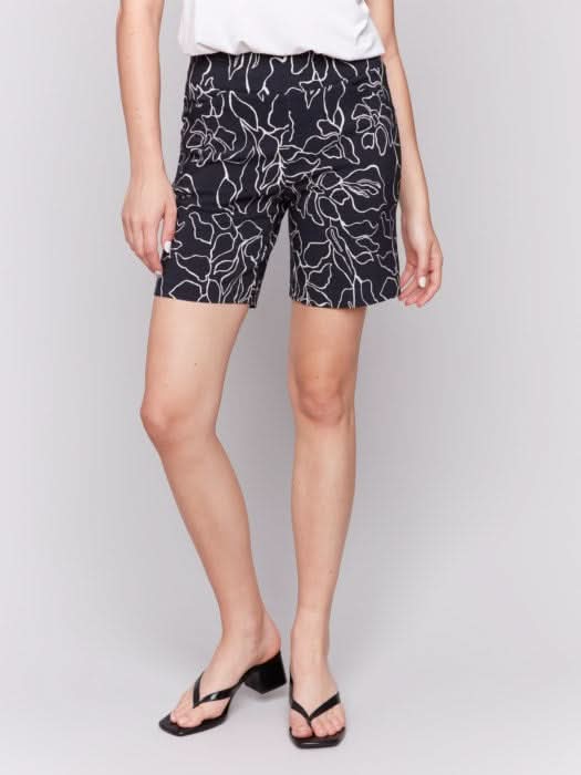 A woman in a white sleeveless top wearing Charlie B's Printed Pull-On Stretch Shorts and black sandals stands with one hand in her pocket.