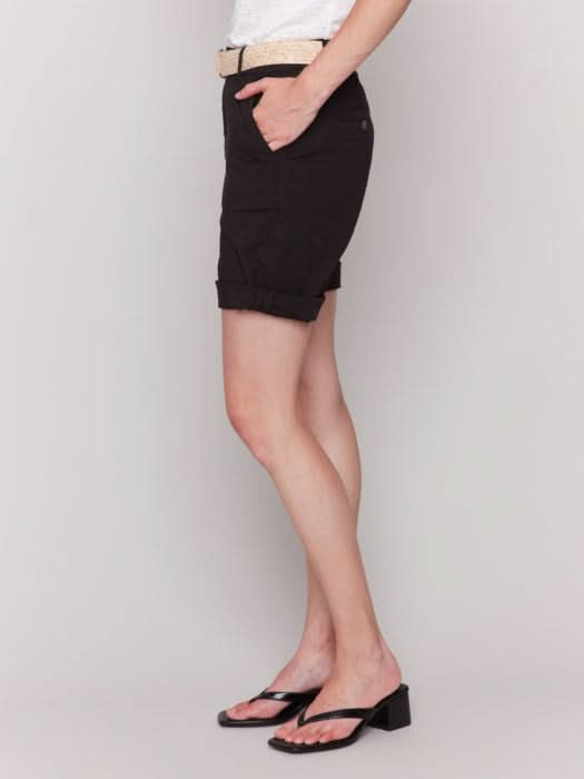 A fashionista against a plain background wears chic Charlie B Cuffed Shorts with a light belt, a crisp white top, and stylish black sandals.