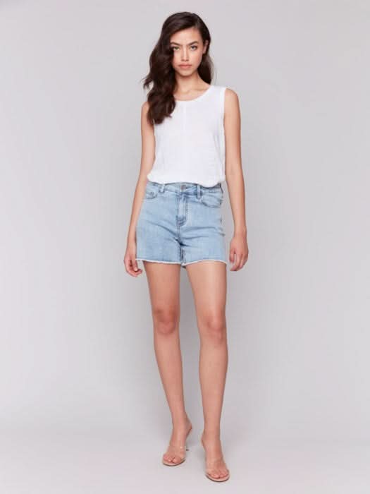 A person wears Charlie B Embellished Stretch Denim Shorts—light blue, embellished, with a frayed hem—paired with a white top and clear high-heeled sandals, standing against a plain light background.