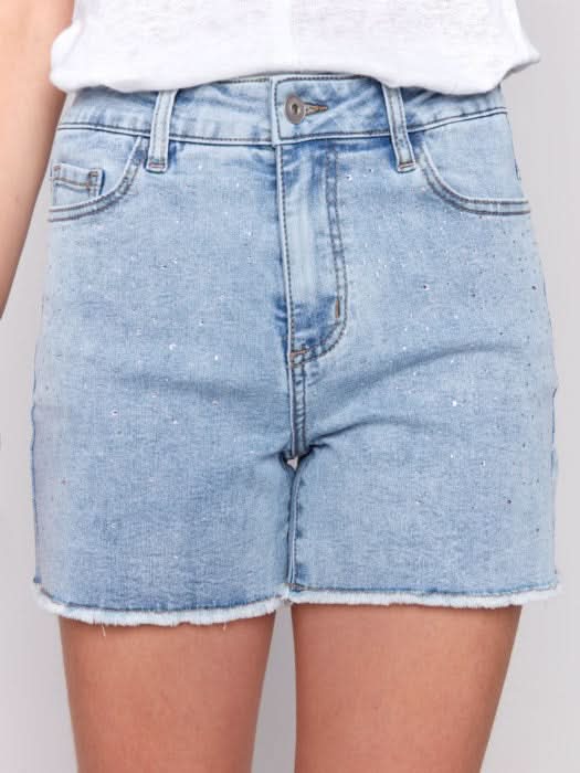 A person wears Charlie B Embellished Stretch Denim Shorts—light blue, embellished, with a frayed hem—paired with a white top and clear high-heeled sandals, standing against a plain light background.