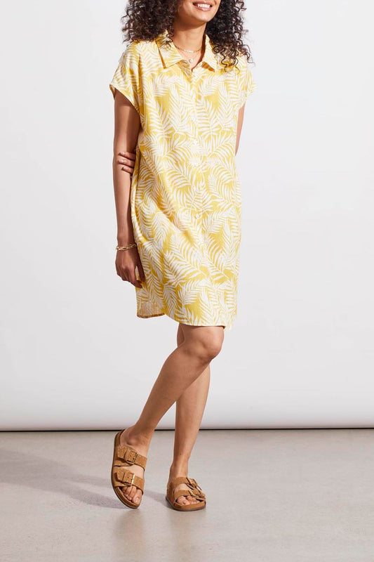 A woman in a stylish Tribal cap sleeve shirt dress with a yellow patterned design and brown sandals standing against a plain background.