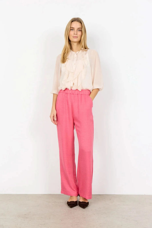 Model wearing Soya Concepts Charissa Pant in pink, styled with a light long-sleeve blouse and dark pointed-toe heels, standing against a plain white wall.