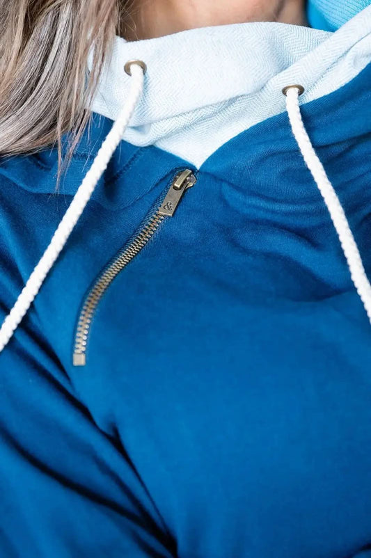 A person wearing an Ampersand Avenue Double Hooded Sweatshirt in a blue cotton blend, featuring a white pocket and drawstrings, stands indoors looking to the side, showcasing a subtle two-tone skyline design.