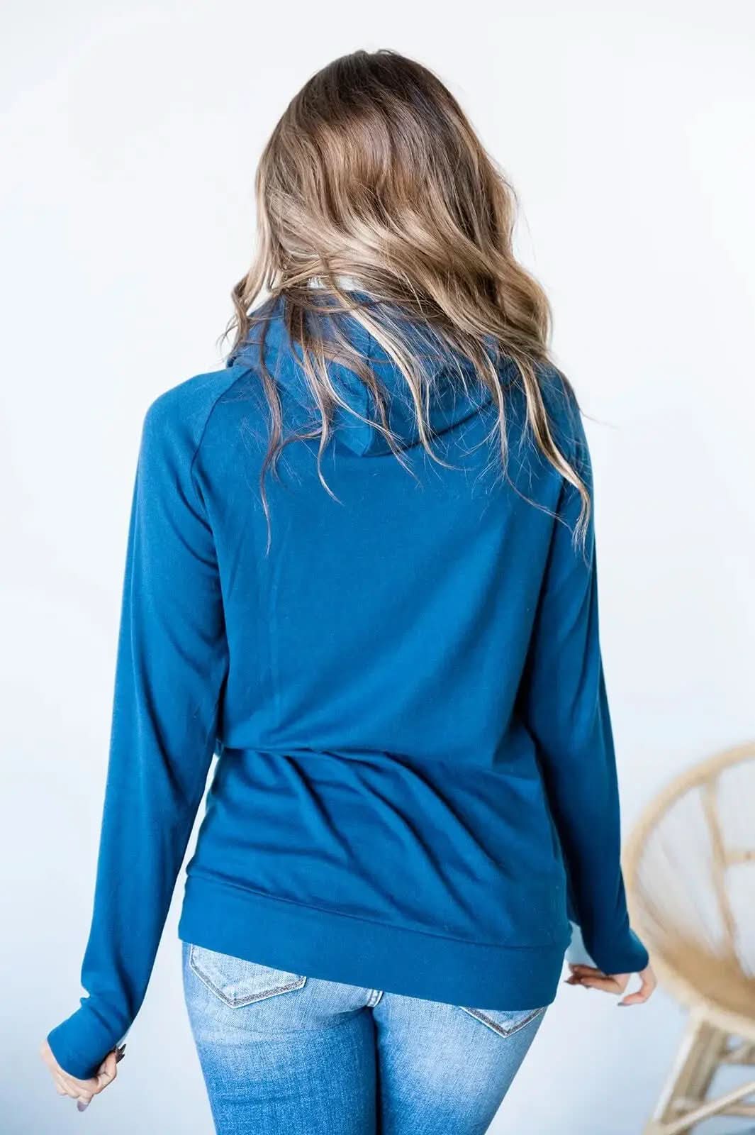 A person wearing an Ampersand Avenue Double Hooded Sweatshirt in a blue cotton blend, featuring a white pocket and drawstrings, stands indoors looking to the side, showcasing a subtle two-tone skyline design.