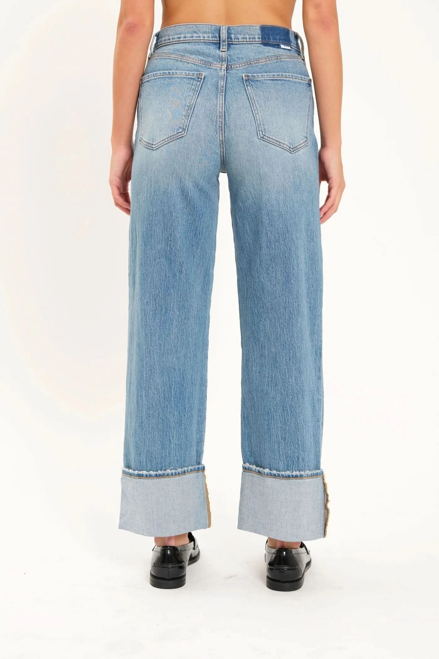 A person is wearing the Eastside High Rise Loose with Wide Cuff jeans in blue, made from organic cotton by Daze, paired with black loafers. The upper body is not shown.