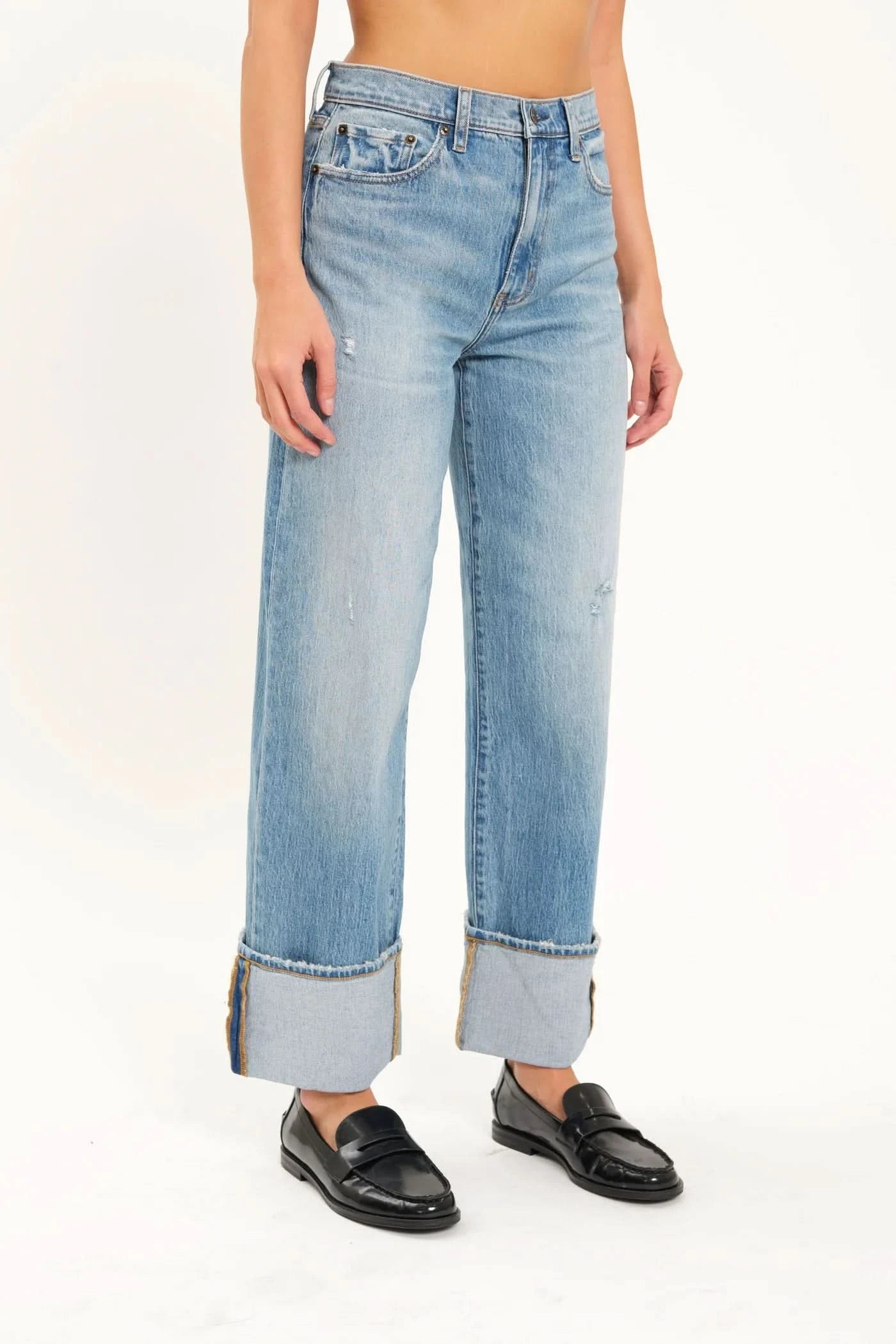 A person is wearing the Eastside High Rise Loose with Wide Cuff jeans in blue, made from organic cotton by Daze, paired with black loafers. The upper body is not shown.
