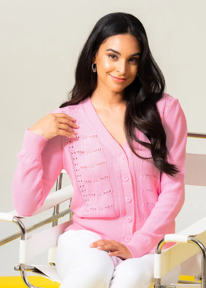 A person seated on a white chair is smiling gently while wearing Elena Wang's stylish and comfortable Front Button Knit Cardigan with a v-neckline and white pants.