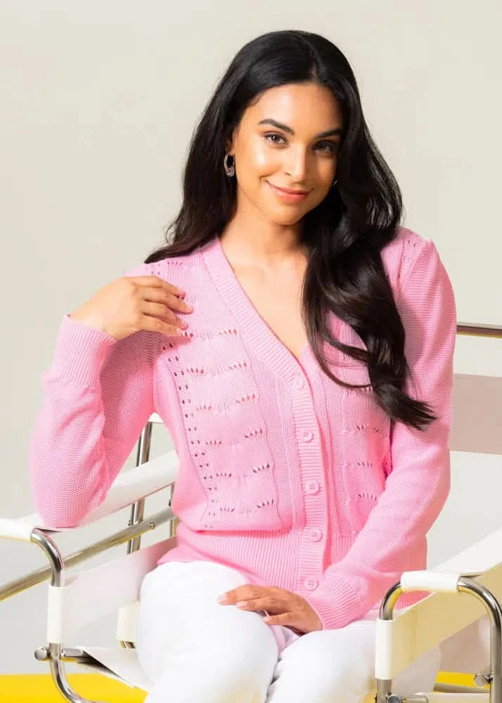 A person seated on a white chair is smiling gently while wearing Elena Wang's stylish and comfortable Front Button Knit Cardigan with a v-neckline and white pants.