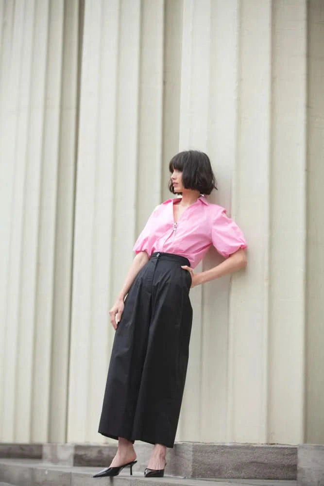 A person poses against a pale background, highlighting the Pink Puffy Blouse by Elena Wang, complemented by white pants and heels, all made from high-quality materials.