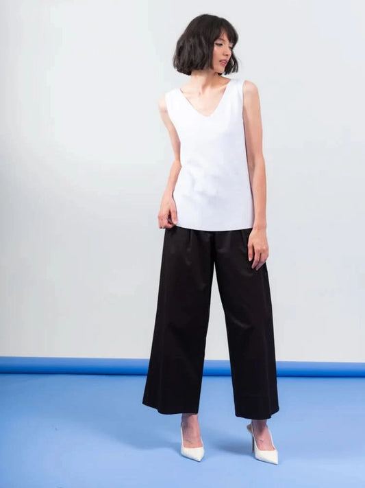The person wears an Elena Wang Reversible Heavy Tank Top and black wide-leg pants with white heels. Posing against a blue-gray background, the fashionable top adds versatility as they look to the side.