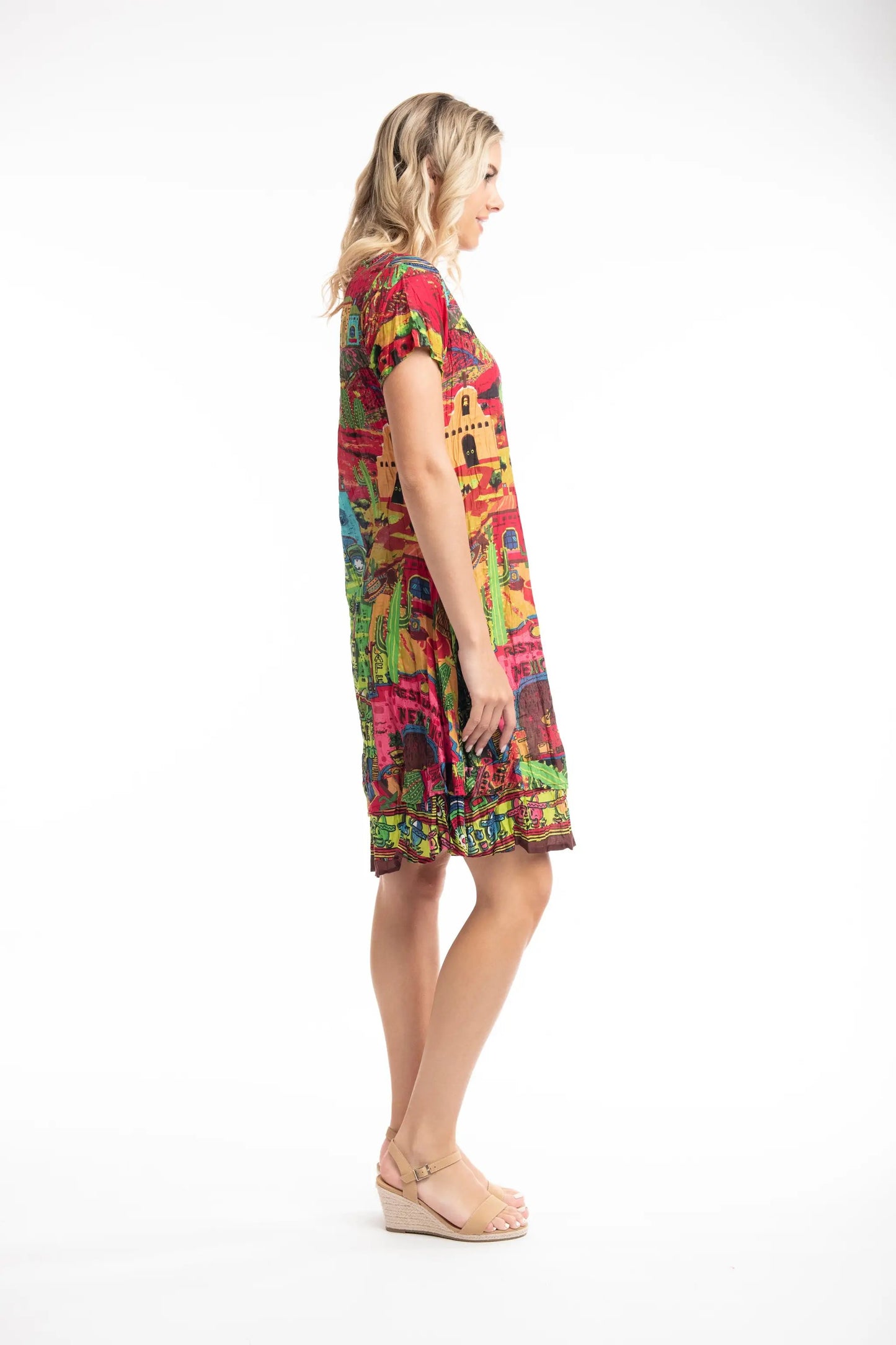 Woman in a colorful printed Orientique El Paso Dress Short slv Bias frill and heels smiling and holding the hem of her dress on a white background.