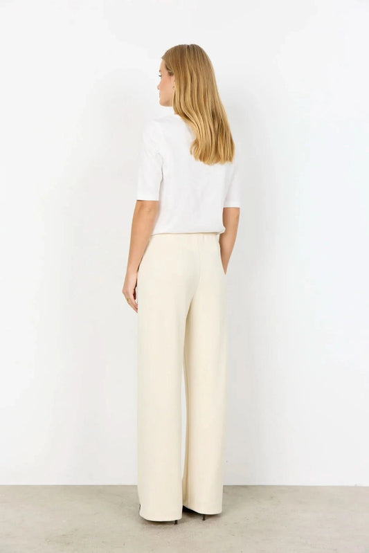 A person wearing Soya Concepts Banu Pant in cream, featuring an adjustable elastic drawstring waist, paired with a white top and brown pointed-toe shoes, stands against a plain background.