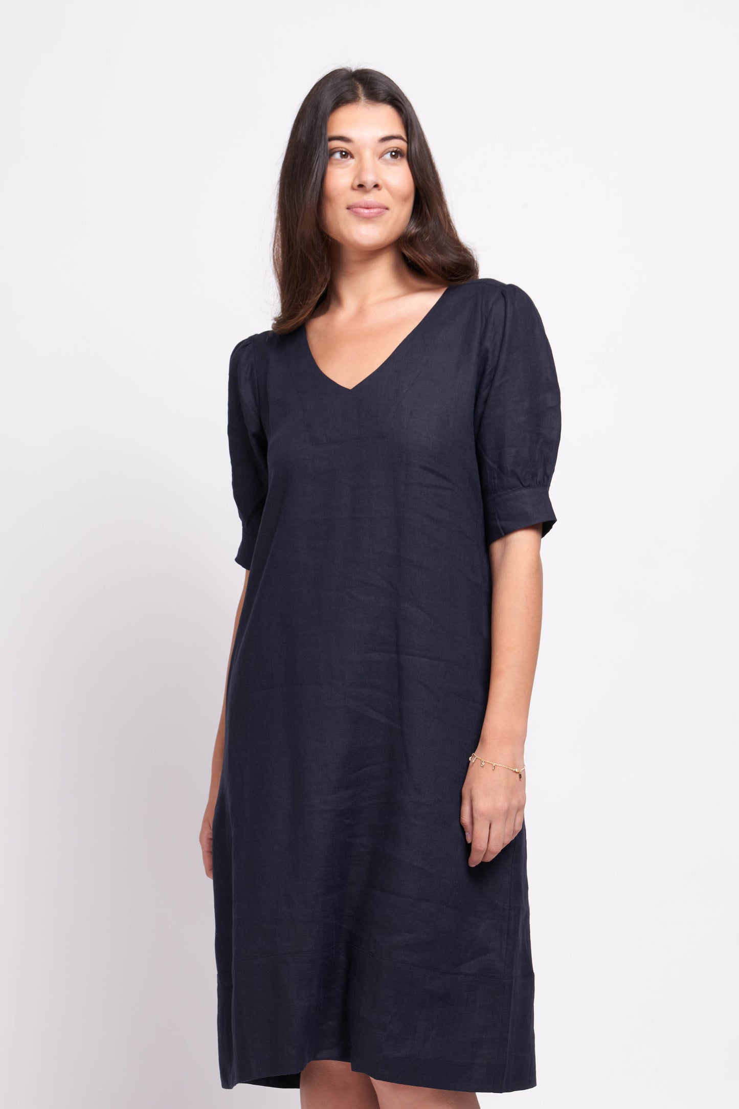 A woman standing in a studio, wearing a Foil navy v-neck linen dress and black sandals, with her hands gently clasped in front of her.