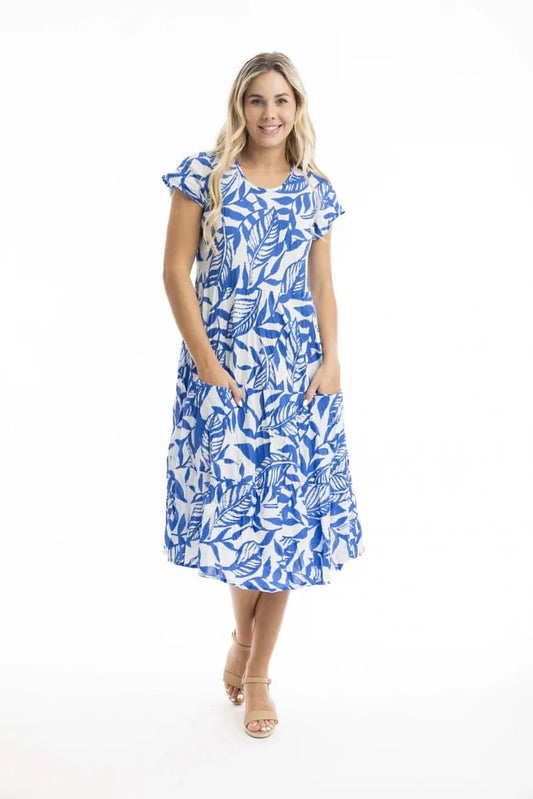 A woman wears the Orientique Bias Blue Dress with Pockets, featuring a blue and white leaf pattern, short sleeves, and playful cut—styled with tan sandals and hands in pockets, set against a plain white background.