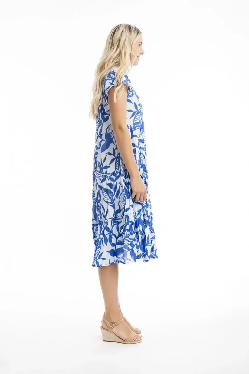 A woman wears the Orientique Bias Blue Dress with Pockets, featuring a blue and white leaf pattern, short sleeves, and playful cut—styled with tan sandals and hands in pockets, set against a plain white background.