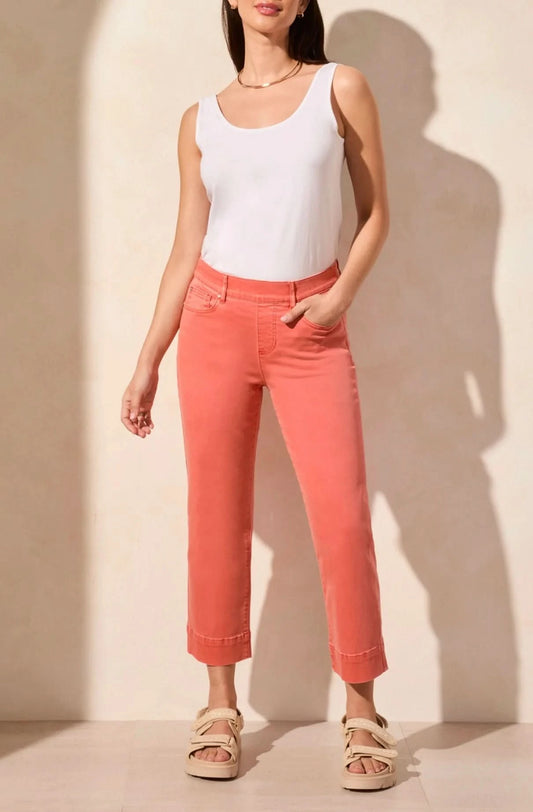 A woman wearing a white sleeveless top and Tribal PULL-ON KICK FLARE CAPRI coral pants with beige sandals stands against a neutral background, exuding comfort with one hand in her pocket.