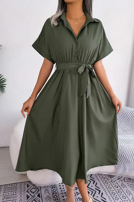 A woman wearing a green, mid-length Loose Fit Button Up Waist Tie Smocked Dress with short sleeves and a waist tie, standing in a stylishly decorated room from Don't Be Chy Boutique.