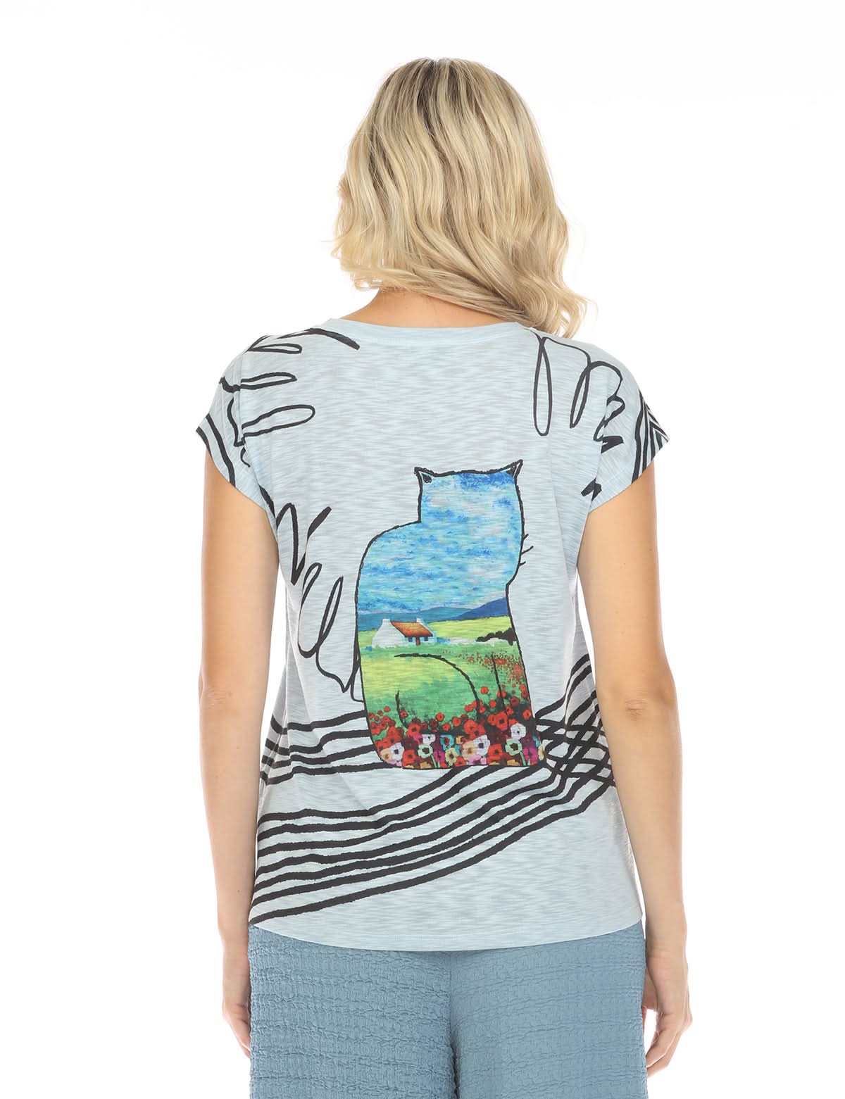 A woman faces away, wearing the Inoah Short Sleeve Crew Neck Top in light blue, featuring a cat silhouette with a house, field, sky, and flowers on the back.