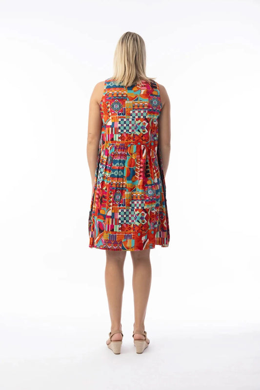 A woman stands on a white background wearing the Orientique Peru Print Dress, a sleeveless, knee-length dress with a colorful retro geometric pattern, paired with tan heeled sandals.