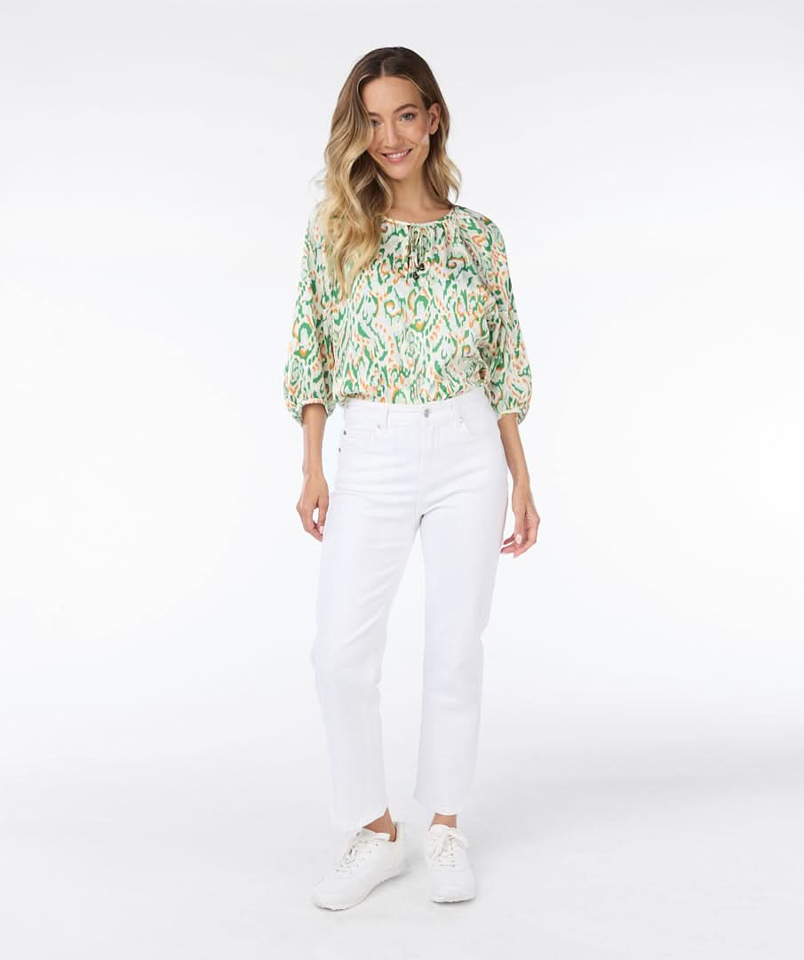 A person wearing an Esqualo Pastel Blouse and white pants stands against a white background, gently touching their hair.