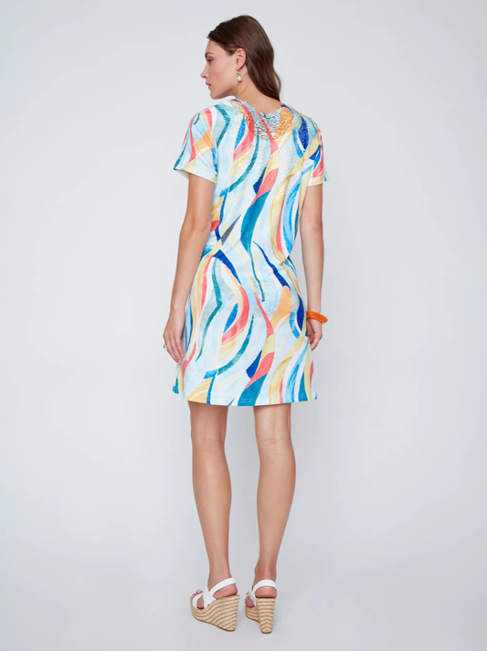 A woman with long brown hair kneels on one knee, wearing CoCo Y Club's Mini Flow Dress—a vibrant, short-sleeved summer dress with an abstract print—paired with earrings and an orange bangle.
