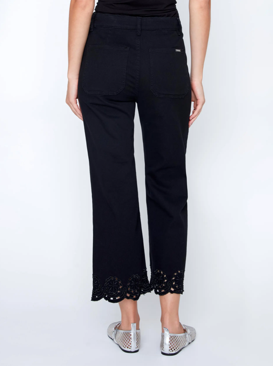 A woman in a black sleeveless top and CoCo Y Club Embroidered Black Trousers holds a silver clutch and wears silver slip-on shoes, standing against a plain white background.