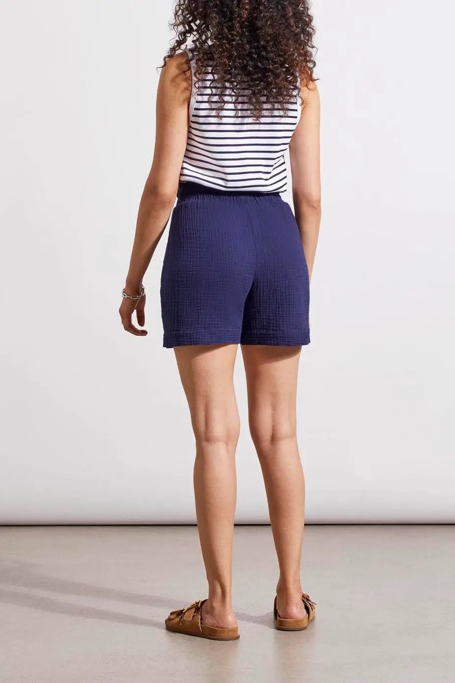 Woman in a casual blue romper with Tribal elastic waistband shorts, wearing a striped undershirt and brown sandals, poses for a fashion display.