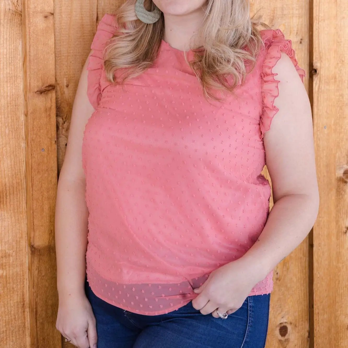 Woman standing, partial view, wearing a pink Don't Be Chy Boutique Swiss Dot Ruffle Sleeveless Top and denim jeans, leaning against a wooden door.