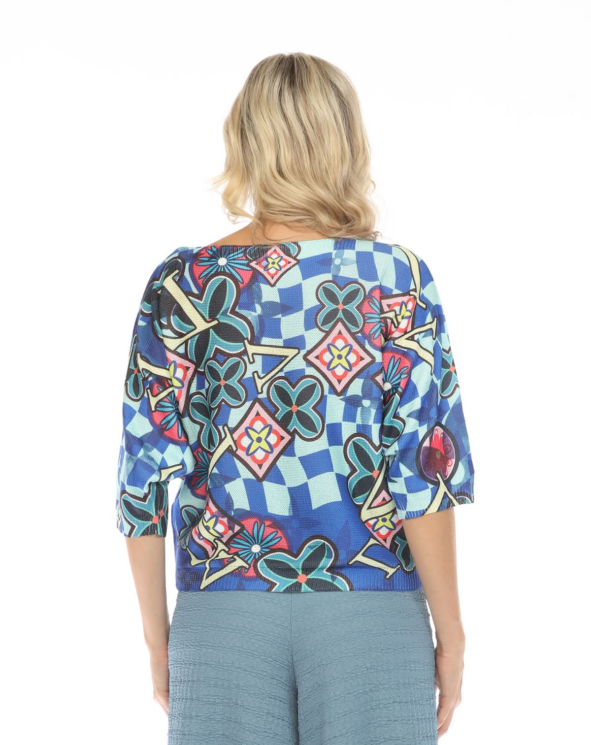 A person with shoulder-length blonde hair is shown from behind, wearing the Inoah 3/4 Sleeve Dolman Sweater featuring a colorful geometric and floral pattern, paired with blue textured pants.