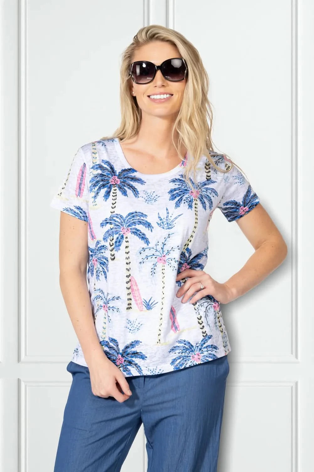A woman wearing sunglasses and blue pants smiles in front of a white panel wall, modeling the Variations Short Sleeve Top with a blue and pink tropical palm tree print.