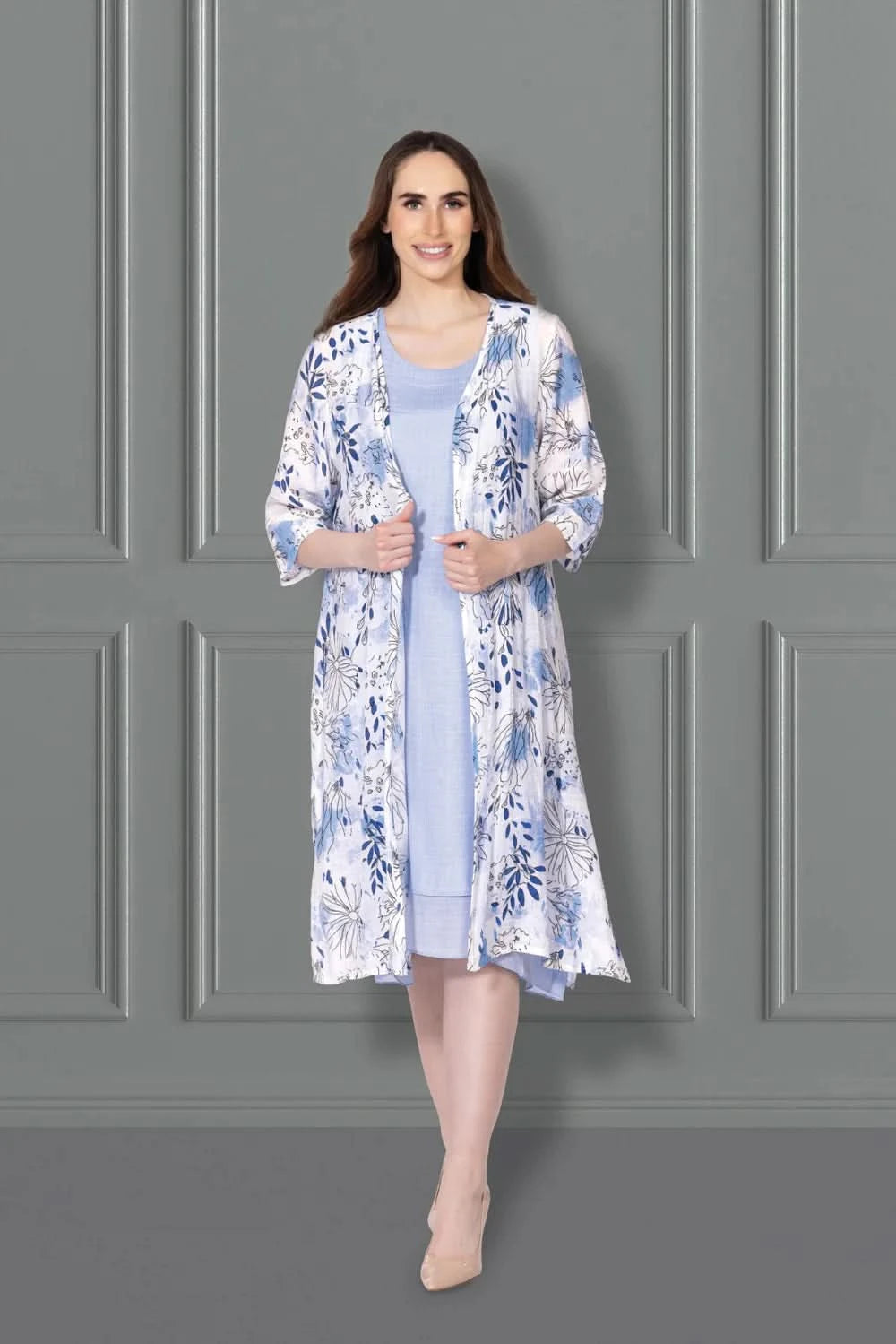 A woman stands against a gray paneled wall, wearing the Variations Solid Dress with Floral Print Jacket Set—a chic two-piece featuring a light blue dress and elegant floral overcoat, paired with beige heels. Perfect for stylish occasions.