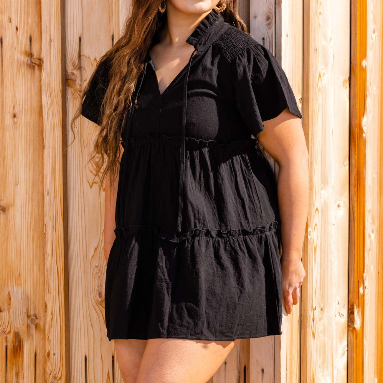 A woman in a black ruffled mini dress with puff sleeves and a tied neckline, holding the skirt, standing outdoors by Don't Be Chy Boutique.