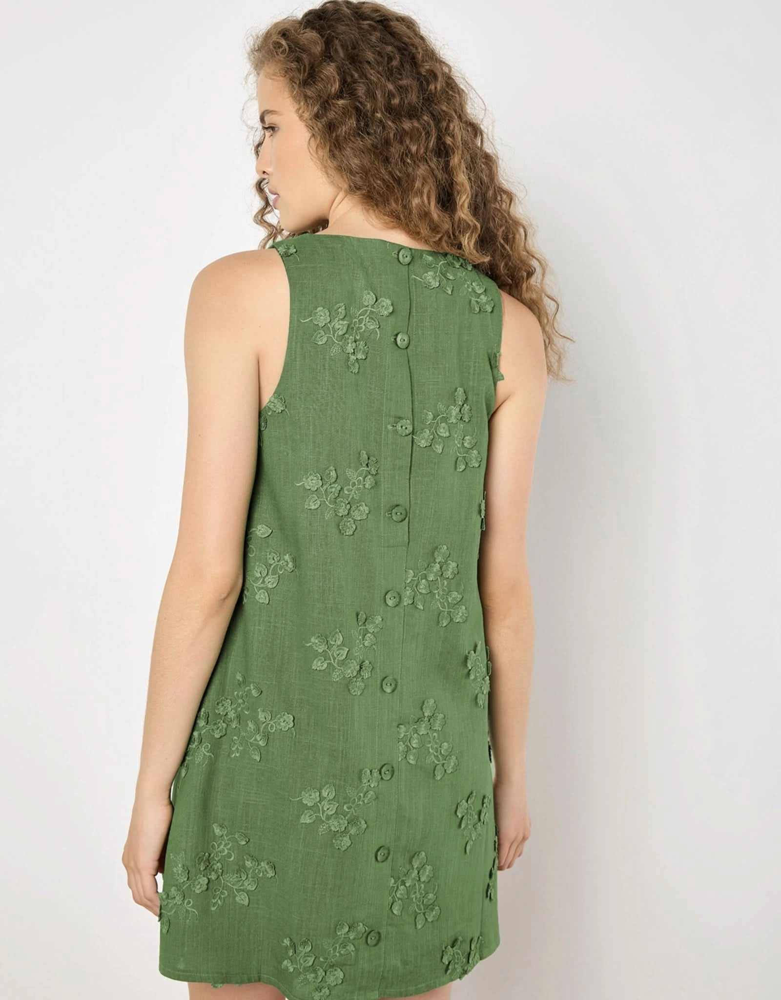 A woman stands by a white wall wearing the Apricot 3D Floral Embroidered Dress in green, styled with black sandals.