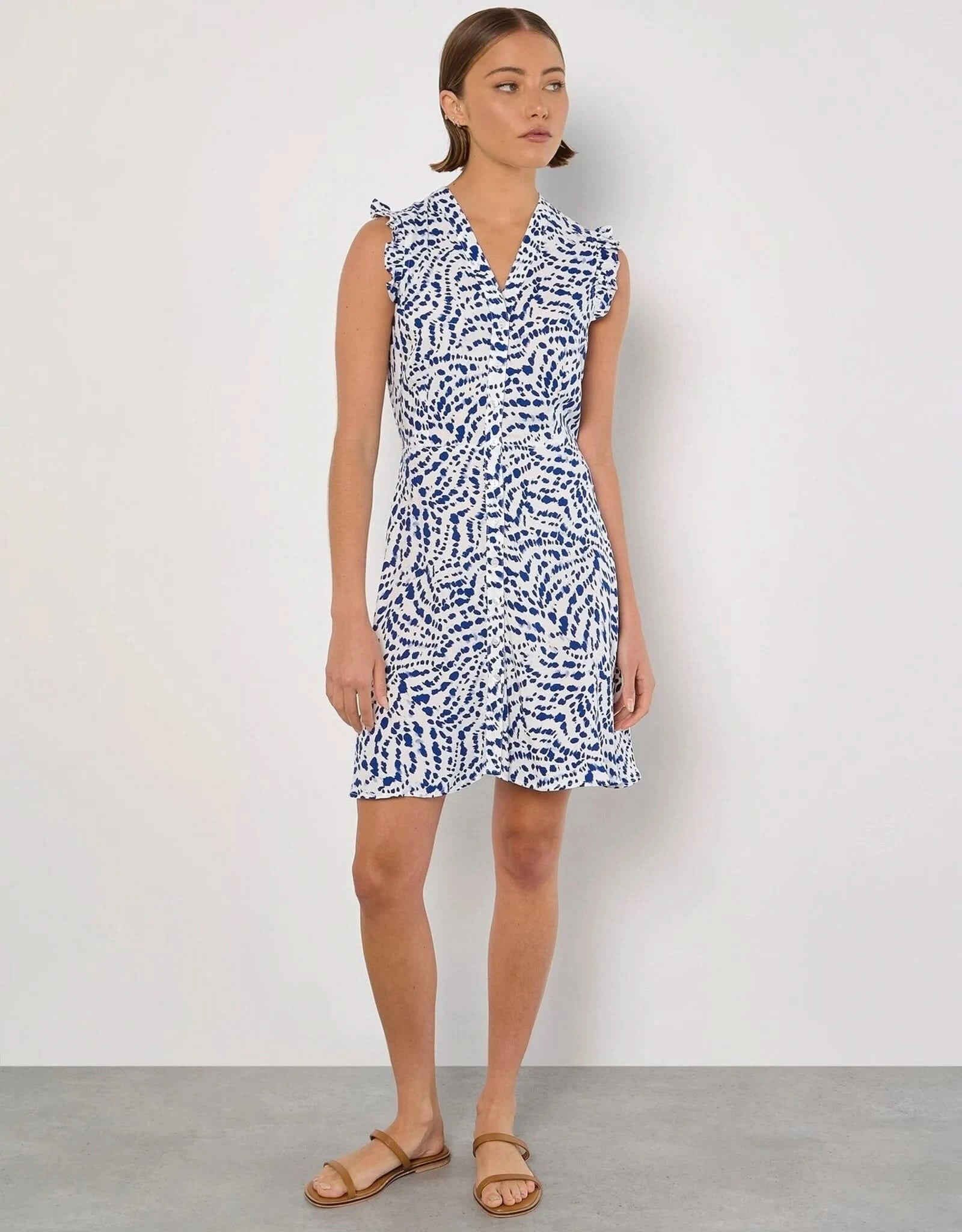 A woman stands against a white background wearing the Apricot Wavy Spot Button Up Dress, ideal for summer with blue abstract dot patterns and ruffled short sleeves.