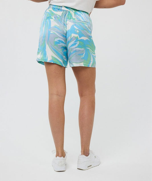 A woman stands on a stone staircase by the water wearing Esqualo's Electric Wave Shorts and matching top, sporting white sandals with one hand on the railing and the other in her pocket.