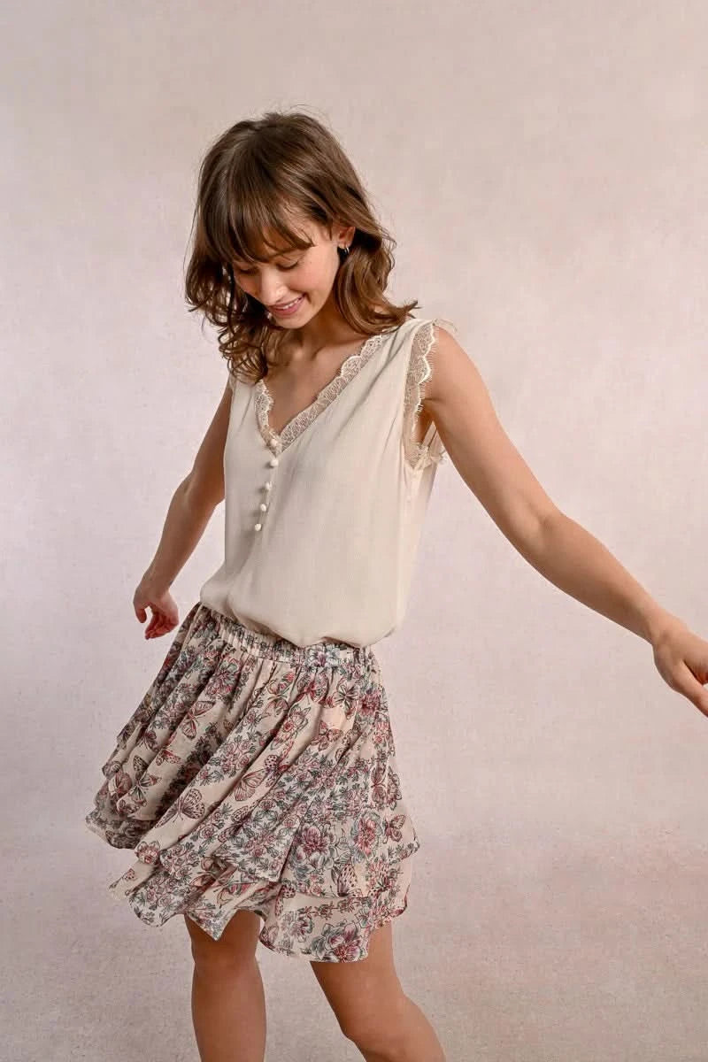 A woman embodies feminine elegance in Molly Bracken's Lace Camisole, a cream sleeveless top with delicate lace trim around the v-neckline and a row of small buttons down the front.