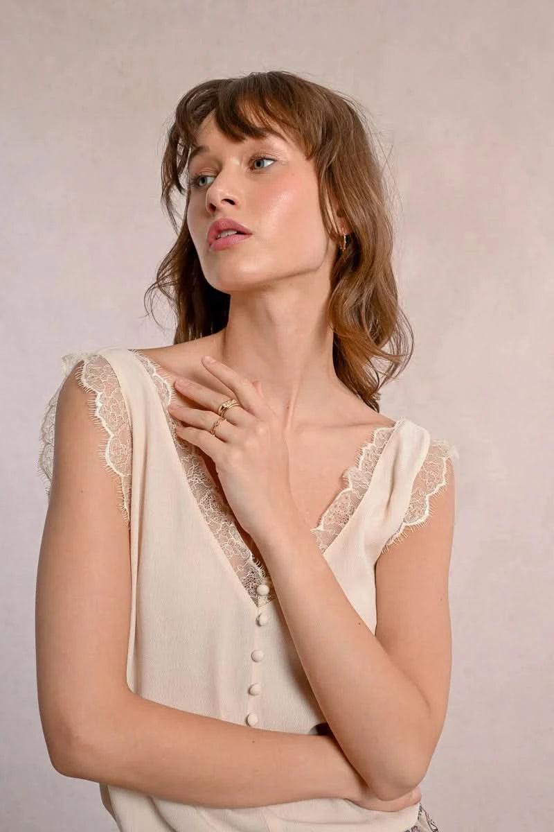 A woman embodies feminine elegance in Molly Bracken's Lace Camisole, a cream sleeveless top with delicate lace trim around the v-neckline and a row of small buttons down the front.