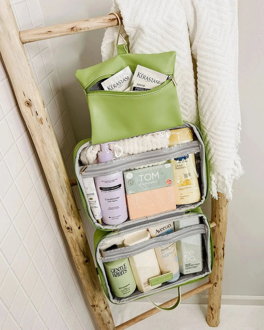 A Louenhide Baby Emma & Audrey Cosmetic Set in green, featuring a front zipper pocket with a partially visible "TO DO" list inside, surrounded by a comb, travel-sized bottle, and white flowers on a tiled surface.