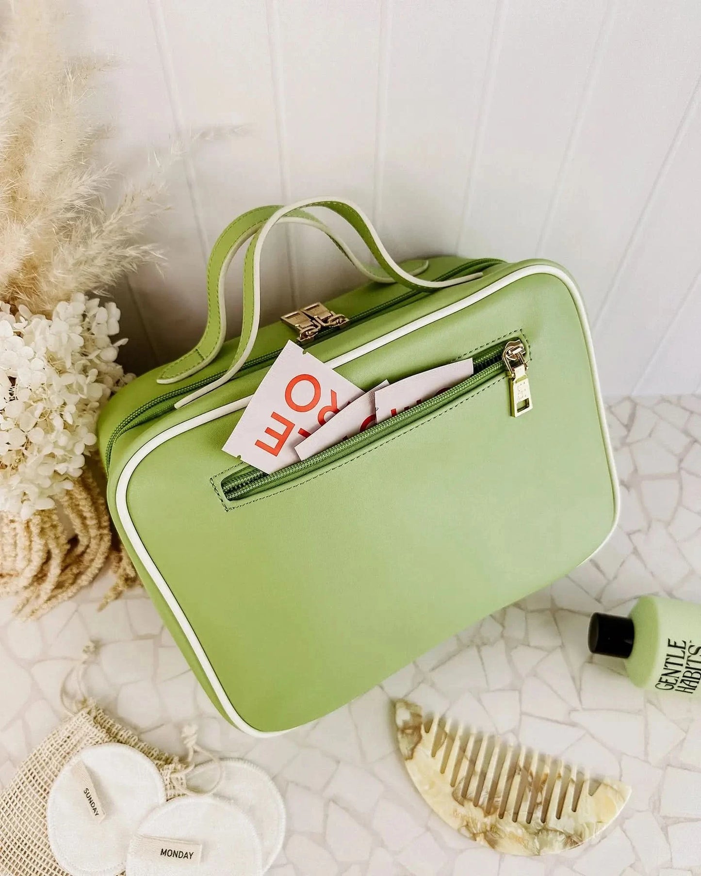 A Louenhide Baby Emma & Audrey Cosmetic Set in green, featuring a front zipper pocket with a partially visible "TO DO" list inside, surrounded by a comb, travel-sized bottle, and white flowers on a tiled surface.