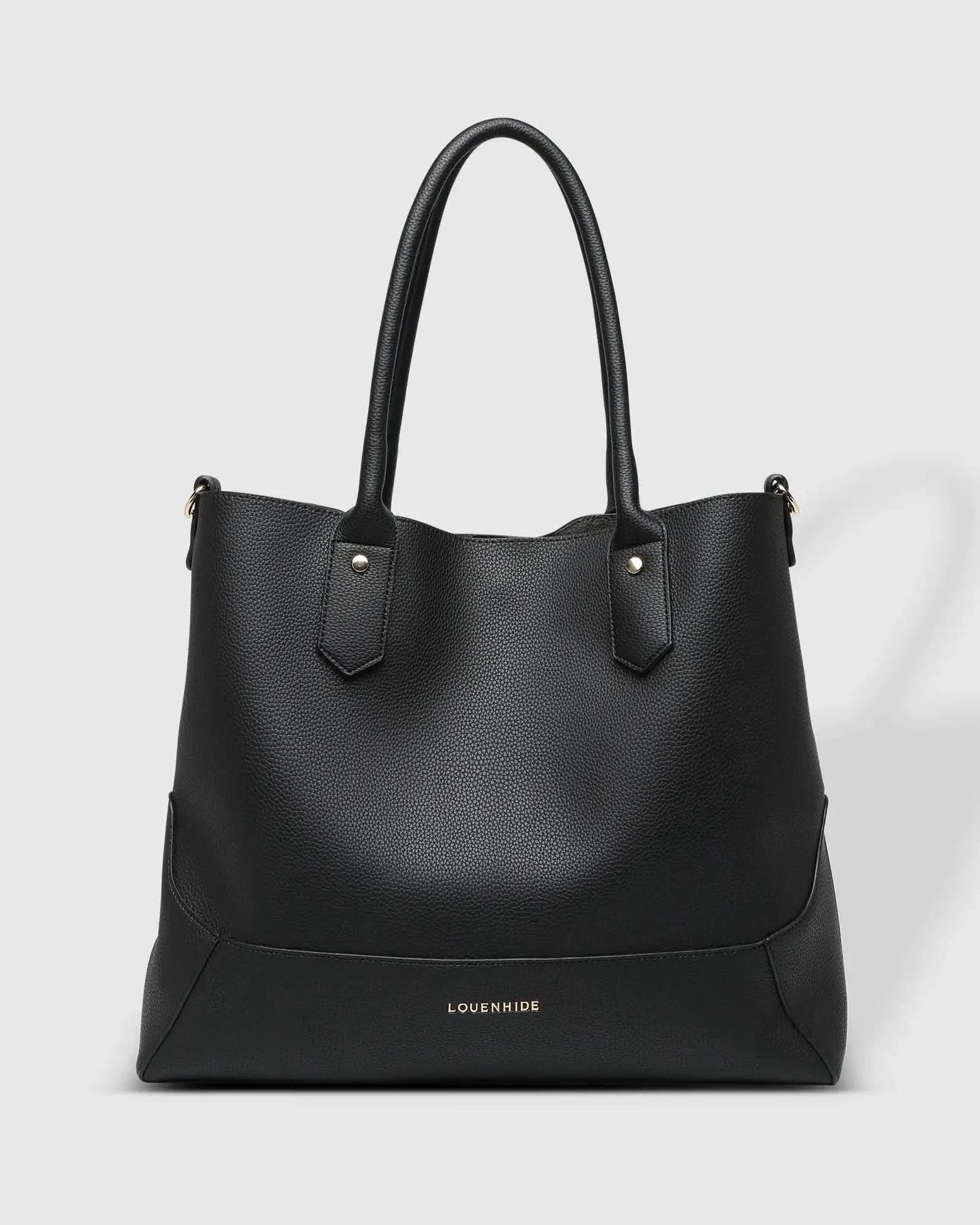 A person with blonde hair in a white outfit poses against a white wall, holding a large black Portsea Tote Bag from Louenhide made of vegan leather over their shoulder.