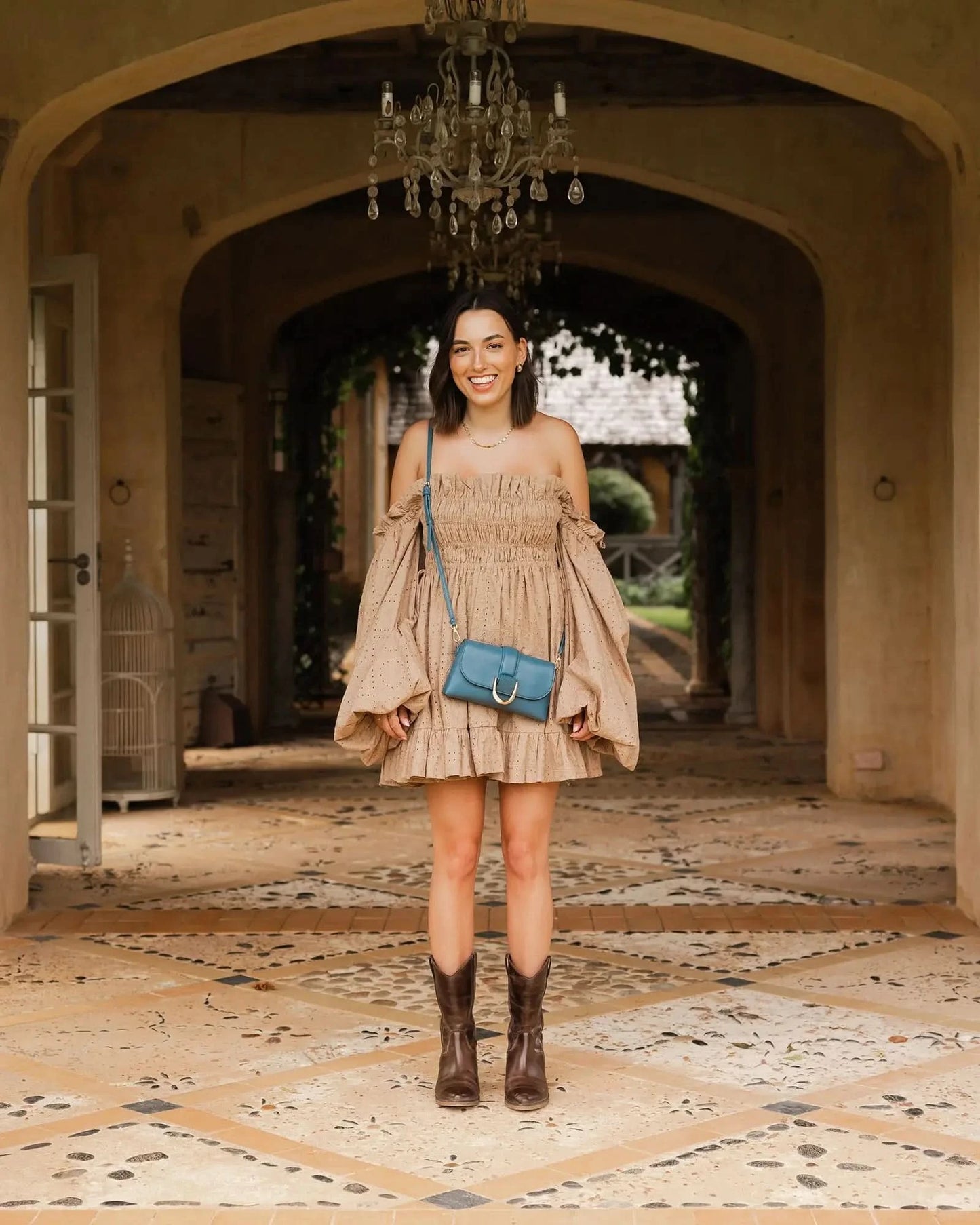 A person wearing a beige off-shoulder dress, brown boots with statement gold hardware buckles, and holding Louenhide's The Pixie Crossbody Bag in blue stands on a stone-tiled floor.