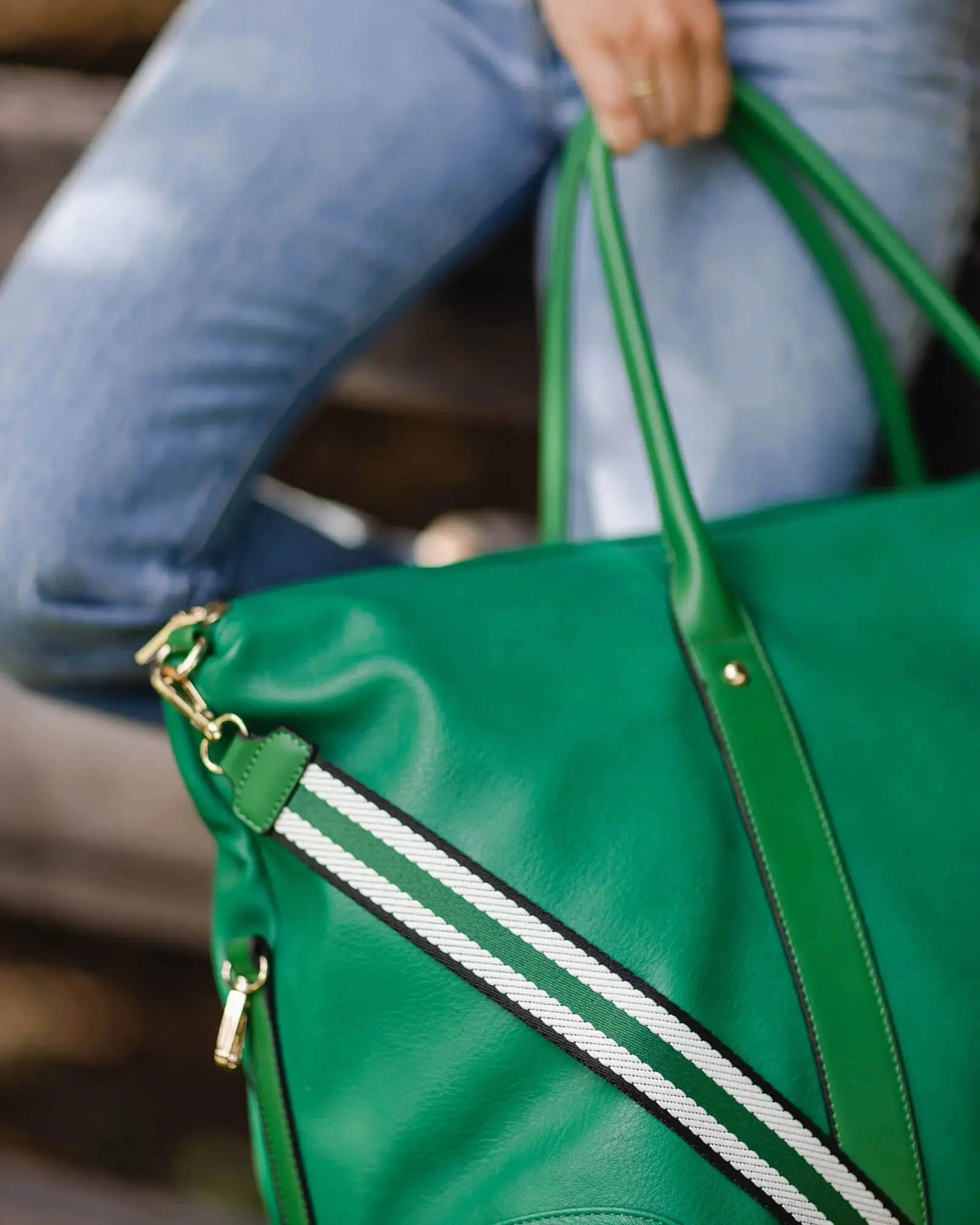 A person holding a large Alexis Stripe Travel Bag by Louenhide, featuring statement stripe webbing and gold hardware, wearing blue jeans.