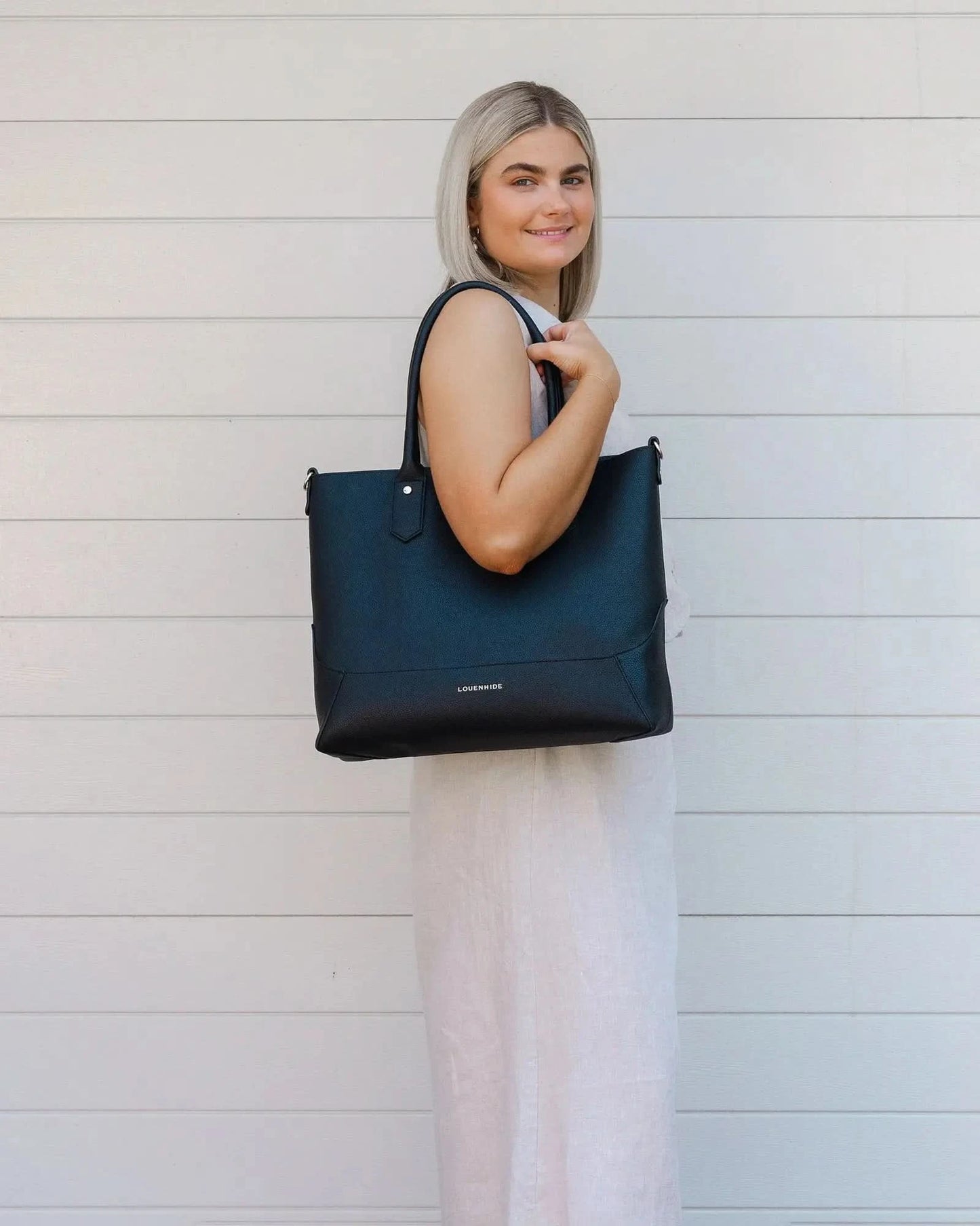 A person with blonde hair in a white outfit poses against a white wall, holding a large black Portsea Tote Bag from Louenhide made of vegan leather over their shoulder.