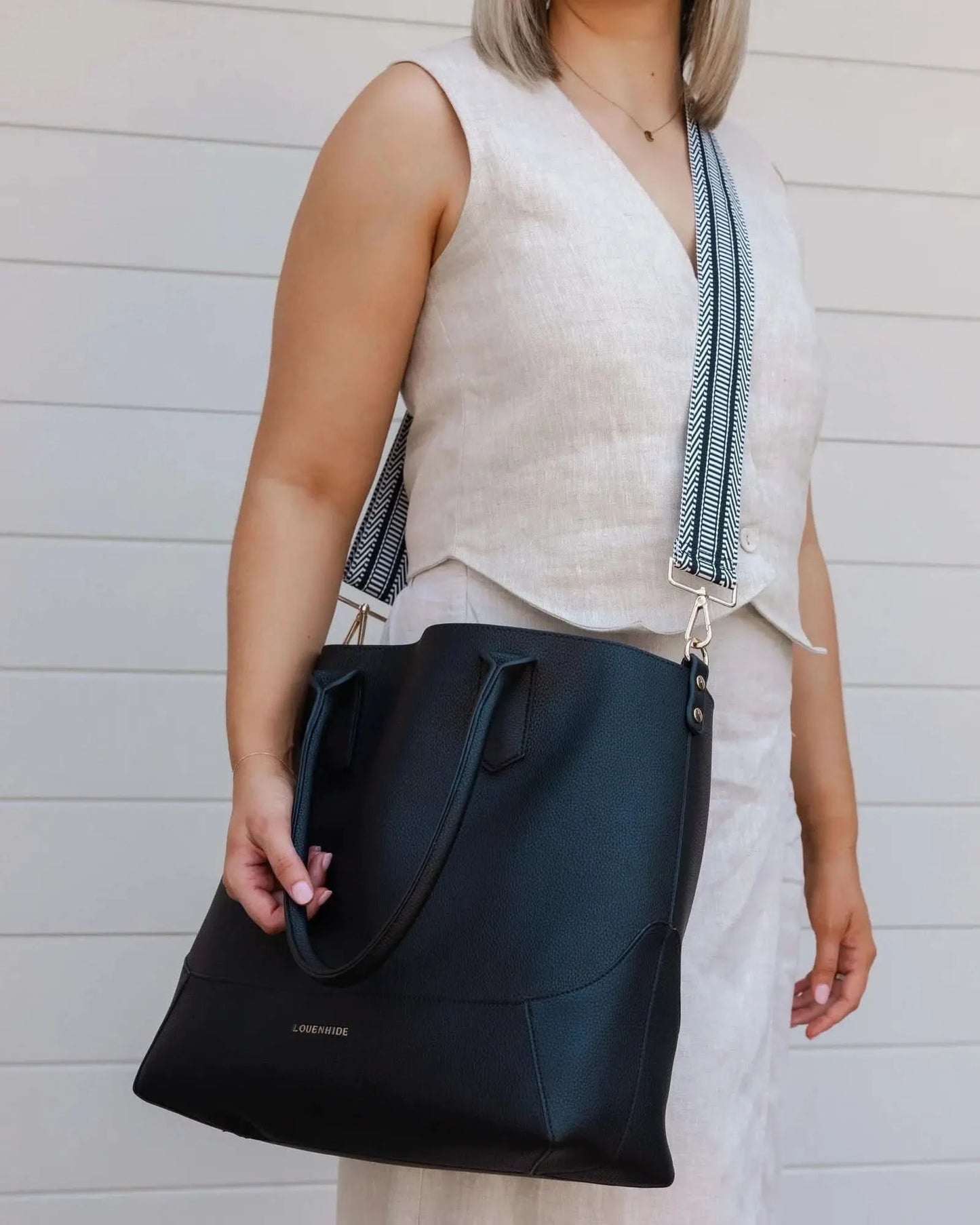 A person with blonde hair in a white outfit poses against a white wall, holding a large black Portsea Tote Bag from Louenhide made of vegan leather over their shoulder.