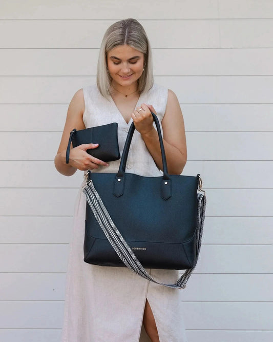 A person with blonde hair in a white outfit poses against a white wall, holding a large black Portsea Tote Bag from Louenhide made of vegan leather over their shoulder.