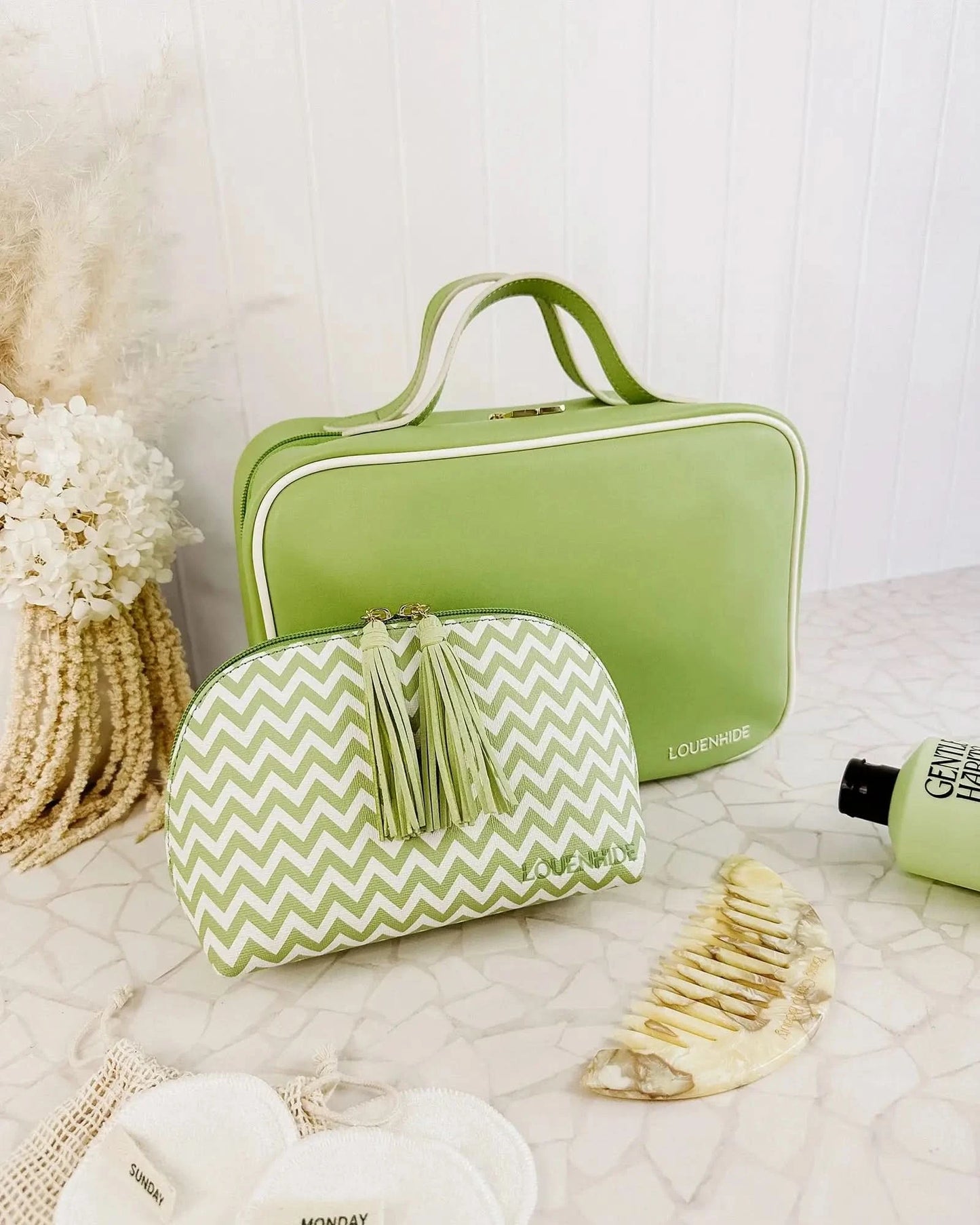 A Louenhide Baby Emma & Audrey Cosmetic Set in green, featuring a front zipper pocket with a partially visible "TO DO" list inside, surrounded by a comb, travel-sized bottle, and white flowers on a tiled surface.