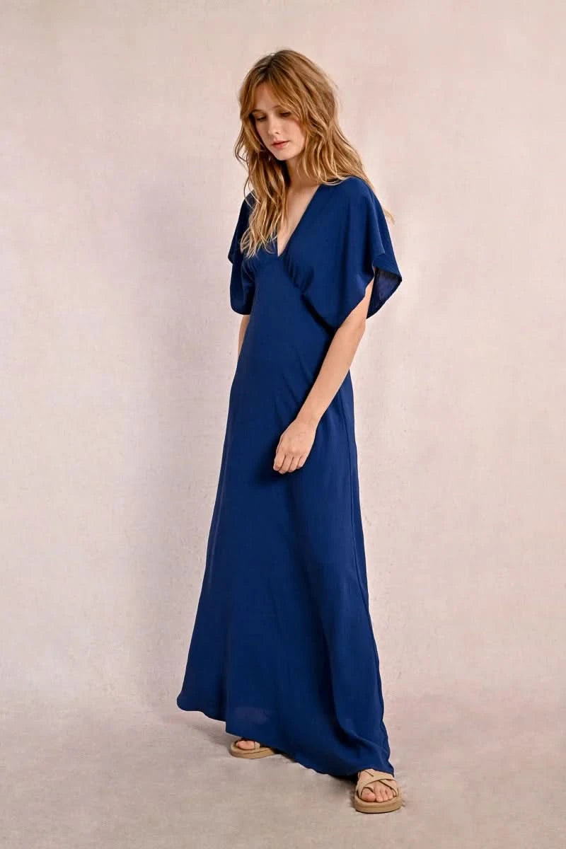 A person elegantly poses against a plain backdrop in a blue Front Pleat Long Marine Dress by Molly Bracken, characterized by its flowy design and short sleeves.